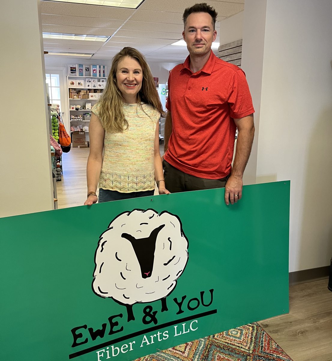 The owners of ewe and You Fiber Arts posing with their sign in the middle of the store.